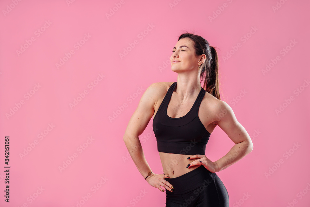 Young pumped up girl coach posing in the studio on a pink background. Dressed in a black top and black pants. Feeling very relaxed and natural, smiles broadly and holds hands on the waist Young pumped up girl coach posing in the studio on a pink background. Dressed in a black top and black pants. Feeling very relaxed and natural, smiles broadly and holds hands on the waist