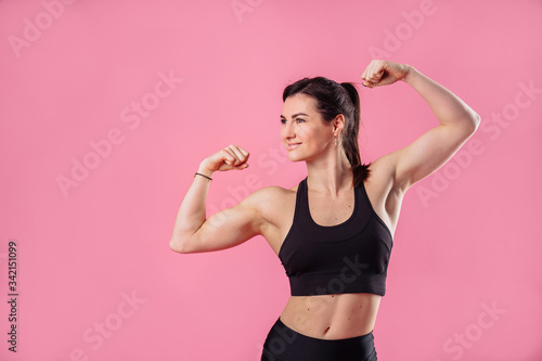 Half lenght portrait of beautiful, pumped up girl in a black tight-fitting top and leggings smiling and showing her big, pumped up muscles in her arms, isolated on pink background in photostudio