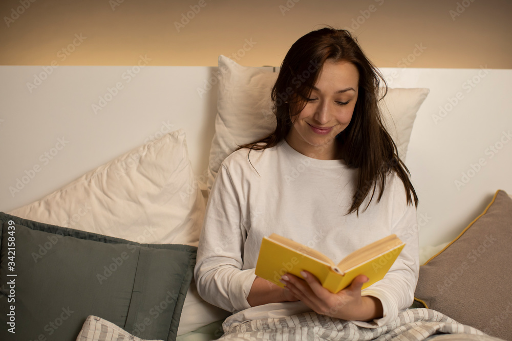 Obraz premium pretty cute dark-haired girl in pajamas smiling while reading book with yellow cover on lazy day modern interior backlighting of bed headboard