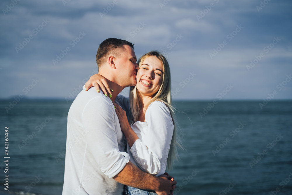 Charming young woman embracing with boyfriend on sea background. Outdoor photo of happy couple standing near ocean. Summer vacation concept. Casual style. Happy together. Outdoor shot Charming young woman embracing with boyfriend on sea background. Outdoor photo of happy couple standing near ocean. Summer vacation concept. Casual style. Happy together. Outdoor shot