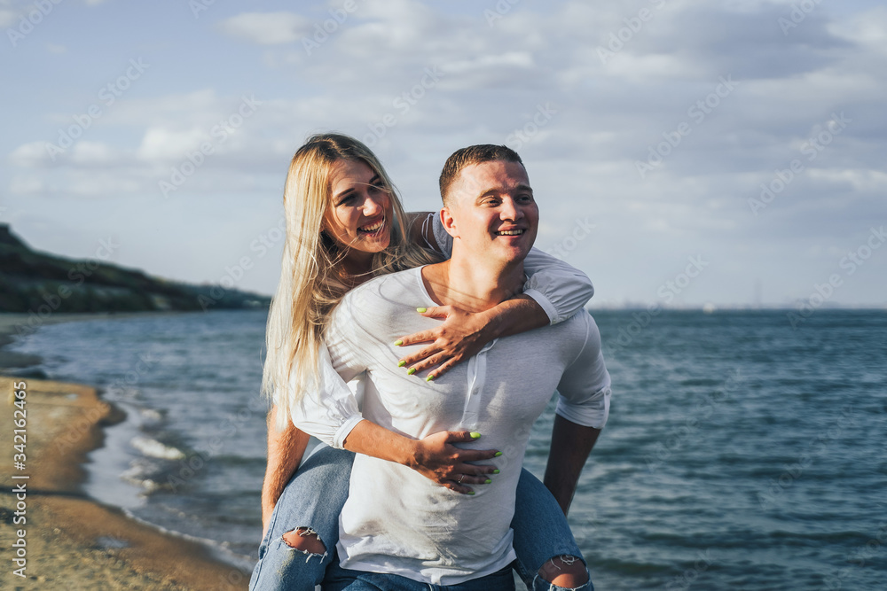 Attractive blonde girl jumped onto her boyfriend's back. He carries her, she laughs and kisses him. They run along the sand on the seashore on a summer evening, lit by the sun. Romantic Date Concept Attractive blonde girl jumped onto her boyfriend's back. He carries her, she laughs and kisses him. They run along the sand on the seashore on a summer evening, lit by the sun. Romantic Date Concept