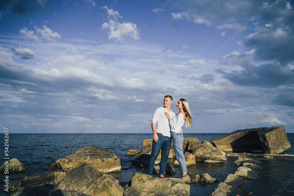 Long shot. Attractive couple in bright clothes standing on stones in the sea against a blue cloudy sky. Lit by the soft evening sun. Looking forward, hug, kissing and enjoying beautiful evening