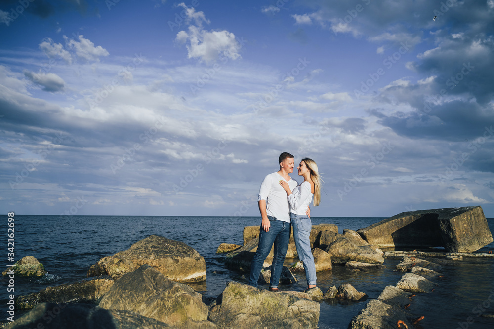 Long shot. Attractive couple in bright clothes standing on stones in the sea against a blue cloudy sky. Lit by the soft evening sun. Looking forward, hug, kissing and enjoying beautiful evening