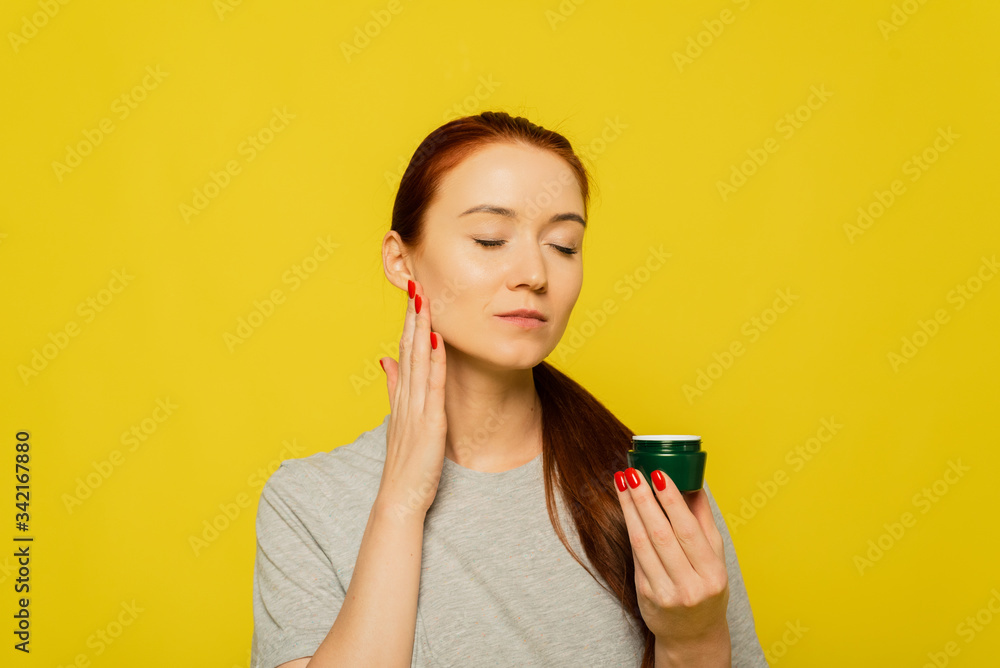A red head girl is applying cream on the face . A girl smears face cream on the yellow background A red head girl is applying cream on the face . A girl smears face cream on the yellow background