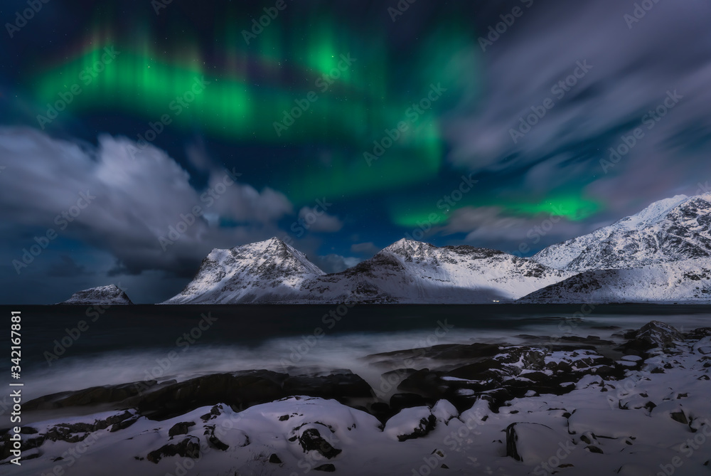 northern lights over the snowcapped mountains on the sea coast, with snow covered rocky beach in the foreground.