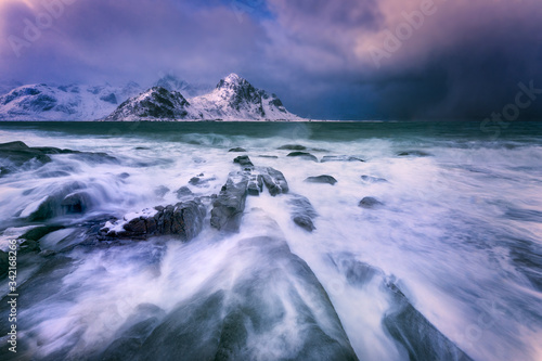 Big waves washing over rocks on the beach with dark storm clouds and snowcapped mountains on the horizon. © Artbotics