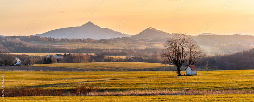 Hilly landscape illuminated by evening sunset. Green grass fields and hills on the horizont. Vivid spring rural countryside. Ralsko Mountain, Czech Republic