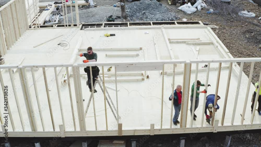 Aerial view of builder standing on steel ladder and nailing planks with manual hammer. Frame house under construction Aerial view of builder standing on steel ladder and nailing planks with manual hammer. Frame house under construction