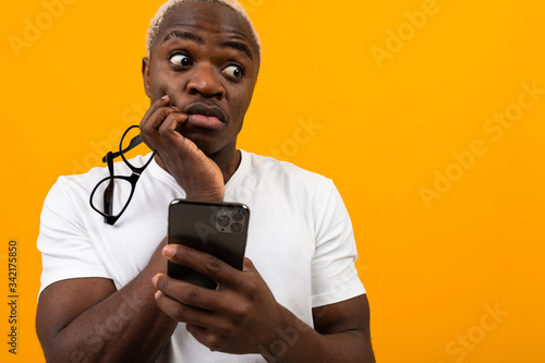 beautiful dark-skinned African with white hair in glasses looks thoughtfully to the side and holds the phone in his hands on a yellow studio background with copy space