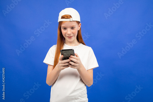 attractive red-haired teenager girl in a white T-shirt communicates in instant messengers holding a smartphone in hand on a blue studio background