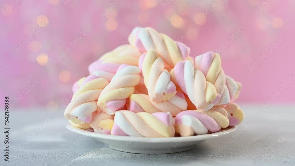 small pile of pastel marshmallows lies on white plate against pink wall with blurry orange flashing lights close view small pile of pastel marshmallows lies on white plate against pink wall with blurry orange flashing lights close view
