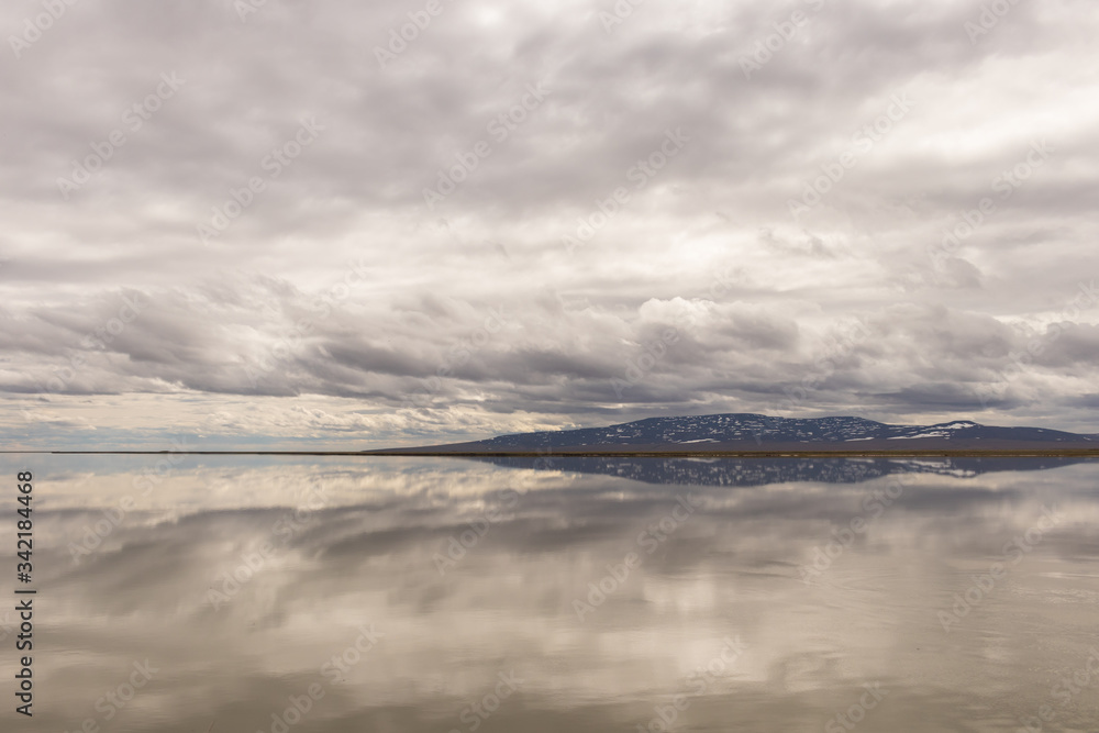 The sloping mountain on the horizon and gray clouds are reflected in the smooth surface of the water on a cloudy day
