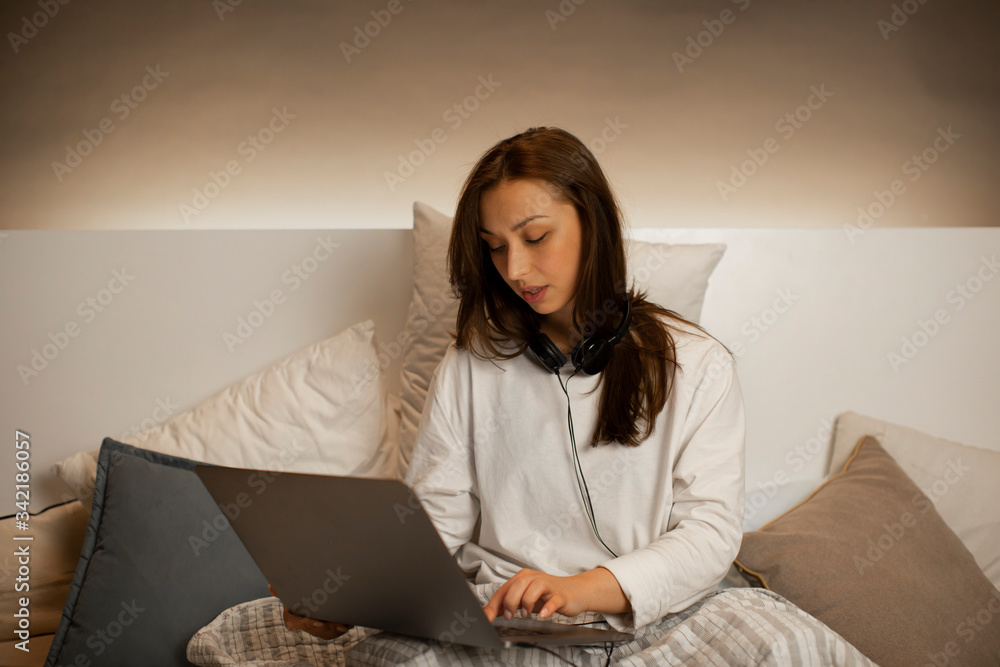Young beautiful sexy girl with long hair in casual wear and headphones sitting in bed, working on laptop from home during self-isolation and quarantine. Home work, remote work concept Young beautiful sexy girl with long hair in casual wear and headphones sitting in bed, working on laptop from home during self-isolation and quarantine. Home work, remote work concept