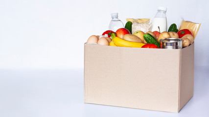 Various grocery items in cardboard box on gray table. Food box with fresh vegetables, fruits, cereals, pasta, milk, eggs and canned goods. Food delivery or donation concept. Copy space.