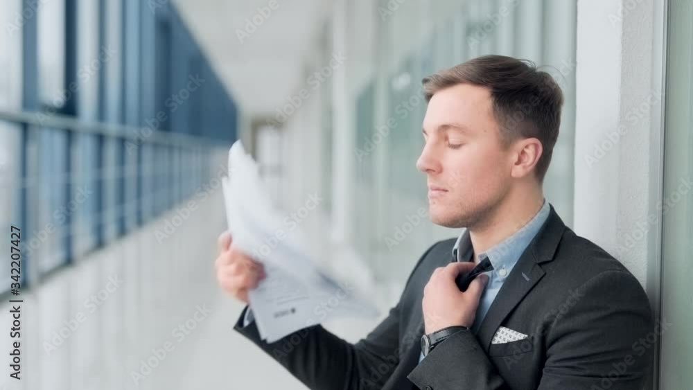 Young businessman sits in hallway and blows fresh air with documents and relaxes tie Young businessman sits in hallway and blows fresh air with documents and relaxes tie