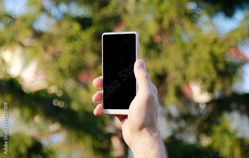 image of man’s hand holding mobile phone with blank screen on background of trees