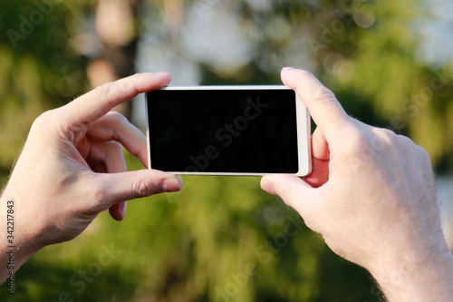 man’s hands holding a white mobile phone with a blank screen on a background of green trees