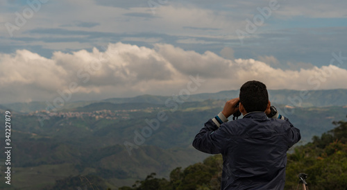 young man looking through binoculars at a mountainous landscape with clouds
