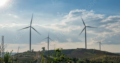 Time lapse Wind turbines on hills landscape with green meadow, hill with flowers, blue cloudy sky with mist.
