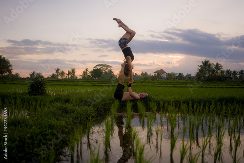 Fototapeta Naklejka Na Ścianę i Meble -  outdoors sunset acroyoga workout - young happy and fit couple practicing acro yoga drill at beautiful rice field enjoying nature and healthy lifestyle doing acrobatic pose
