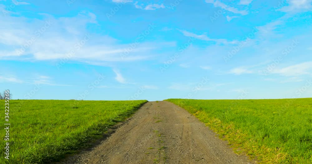 Road gravel stony between two green fields between villages leading to hill with blue sky in the background and moving greenery in strong wind during sunny day captured 4k 60fps Road gravel stony between two green fields between villages leading to hill with blue sky in the background and moving greenery in strong wind during sunny day captured 4k 60fps