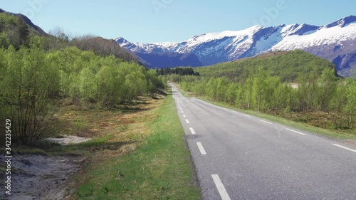 A road stretches through the valley, low forest covering the hills, snow-covered mountain peaks towering on the horizon. The blue cloudless sky above. photo