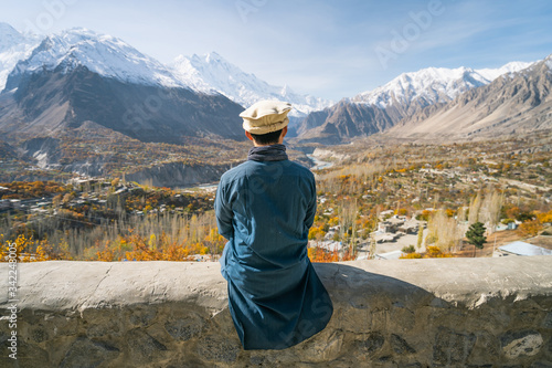 A man with traditional dress sitting on wall and looking at Hunza valley in autumn season, Gilgit Baltistan in Pakistan