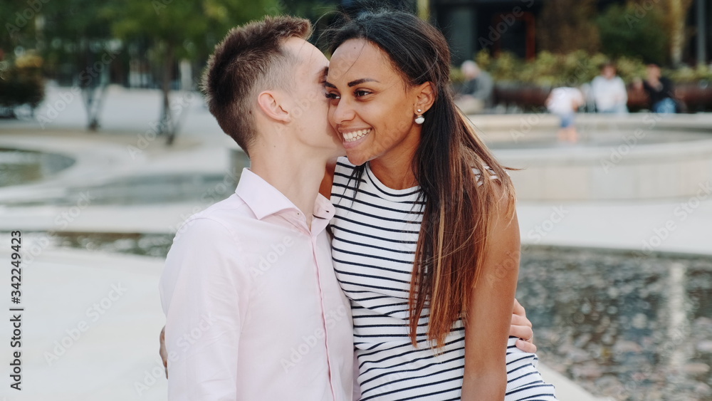 Close-up of young man whispering something in girlfriend's ear sitting on a bench outdoors. They smiling, hugging and talking to each other.