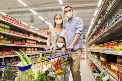 sale, family and pandemic concept - happy mother, father and daughter wearing face protective medical masks for protection from virus disease with shopping cart buying food at supermarket