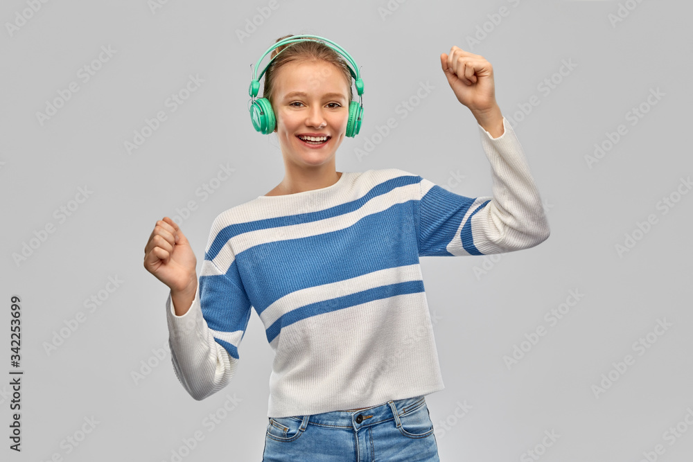 Fototapeta premium music, fun and people concept - happy smiling teenage girl in headphones and pullover dancing over grey background