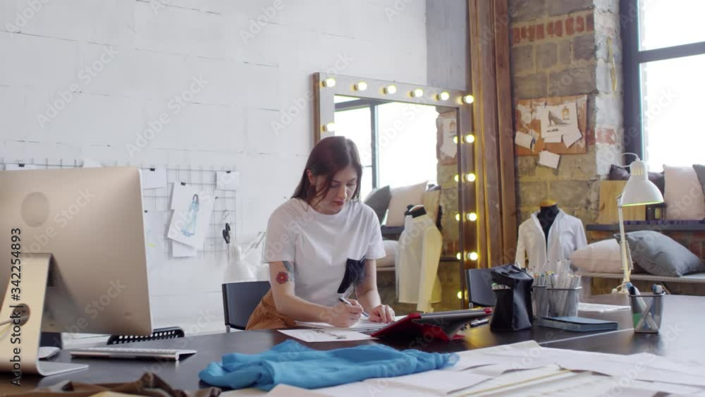 Zoom in of beautiful female designer sitting at worktable in sewing studio and drawing fashion sketch from picture on digital tablet Zoom in of beautiful female designer sitting at worktable in sewing studio and drawing fashion sketch from picture on digital tablet