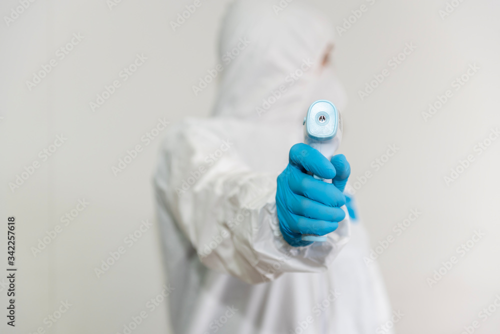Young woman in a protective suit and blue gloves with an infrared thermometer. The girl measures the temperature towards the camera on a white background Young woman in a protective suit and blue gloves with an infrared thermometer. The girl measures the temperature towards the camera on a white background