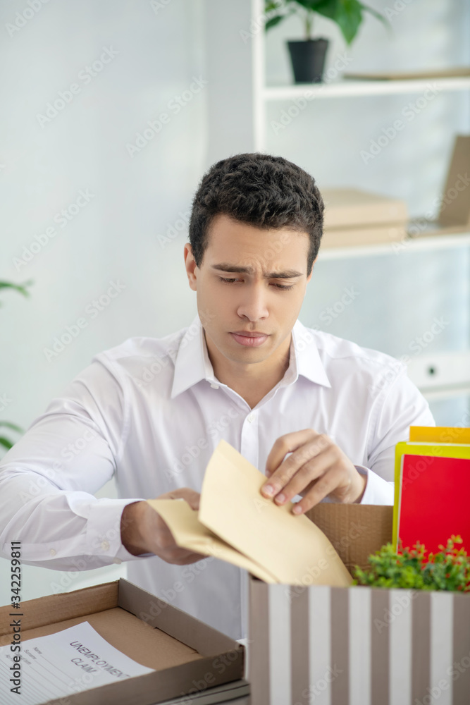 Upset brunette male sitting at his desk, putting envelopes into the box with his personal stuff Upset brunette male sitting at his desk, putting envelopes into the box with his personal stuff