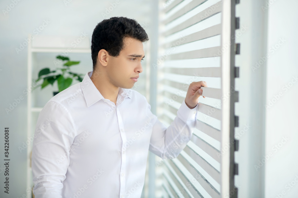 Upset young brunette male standing at the window, touching blinds Upset young brunette male standing at the window, touching blinds