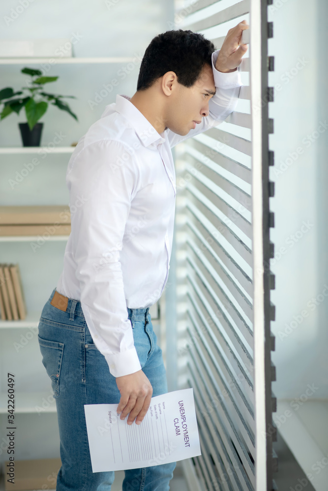 Upset young brunette male standing at the window, leaning his arm and head on blinds, holding unemployment claim Upset young brunette male standing at the window, leaning his arm and head on blinds, holding unemployment claim