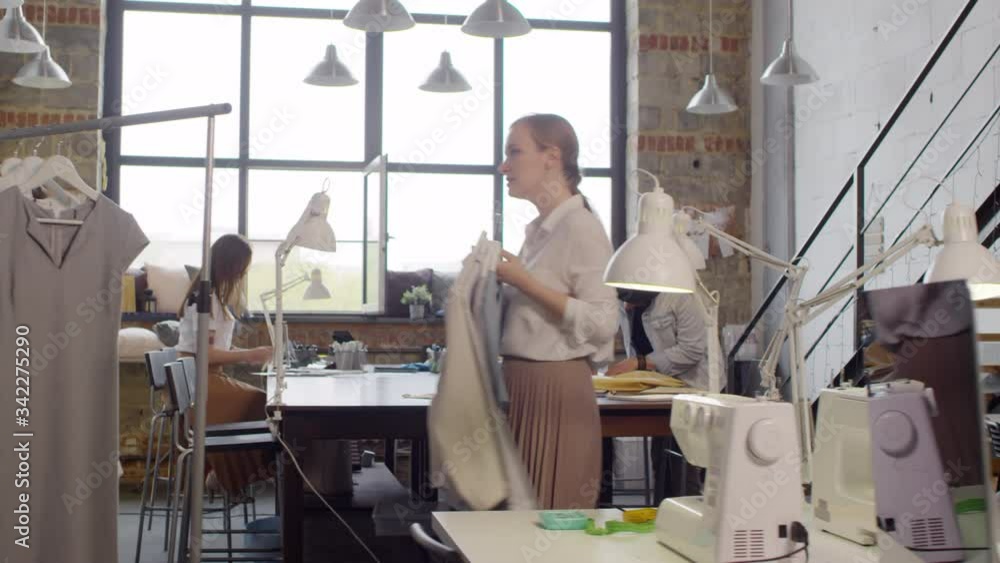 Female fashion designer bringing garments and hanging them on clothing rack while working with colleagues in sewing studio Female fashion designer bringing garments and hanging them on clothing rack while working with colleagues in sewing studio