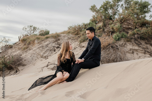 Attractive couple sitting on the sand posing holding hands Black formal shirt and pants