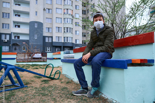 teen boy sits on a bench on playground near high-rise buildings with apartments, a medical mask on his face protects against viruses and dust