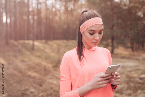 Girl inserts a post on her social media acount after workout. Attractive woman checks achievements after her exercise on smart phone. Young athletic slim brunette sexy runner in pink  running blouse. photo