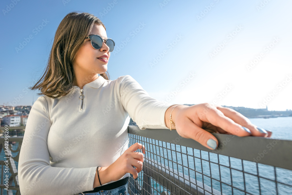Sexy brunette girl is posing with her hands on the fence and turned  towards the sea. Glamorous fashionable trendy background image.