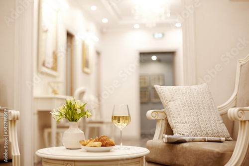A glass of white wine, crispy croissants and a vase of white flowers sit on the table in the cozy living room © Sviatlana