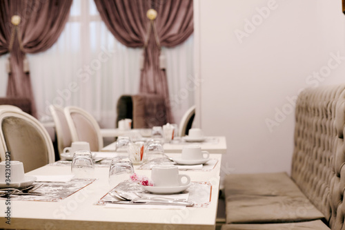 The table is served for Breakfast in a cozy bright dining room with large Windows and dark curtains. On the table are ceramic cups  glasses and Cutlery