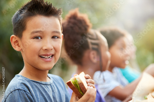 Portrait Of Boy With Friends Eating Healthy Picnic At Outdoor Table In Countryside