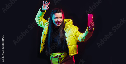 Young girl blogger posing with smart phone in her hands, making selfie on black background and broadcasts live with subscribers and followers. Advertisement concept. Studio shots, copy space for text
