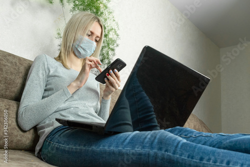 Young blonde woman in a medical mask with a laptop on her lap and a mobile phone in her hands is sitting on the sofa. Concept of work at home, self-isolation, pandemics. © yaroslav1986