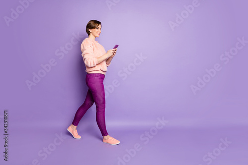 I'm on my way. Full length photo of pretty lady holding telephone chatting with friends walking down street wear casual stylish pink sweater trousers isolated purple color background