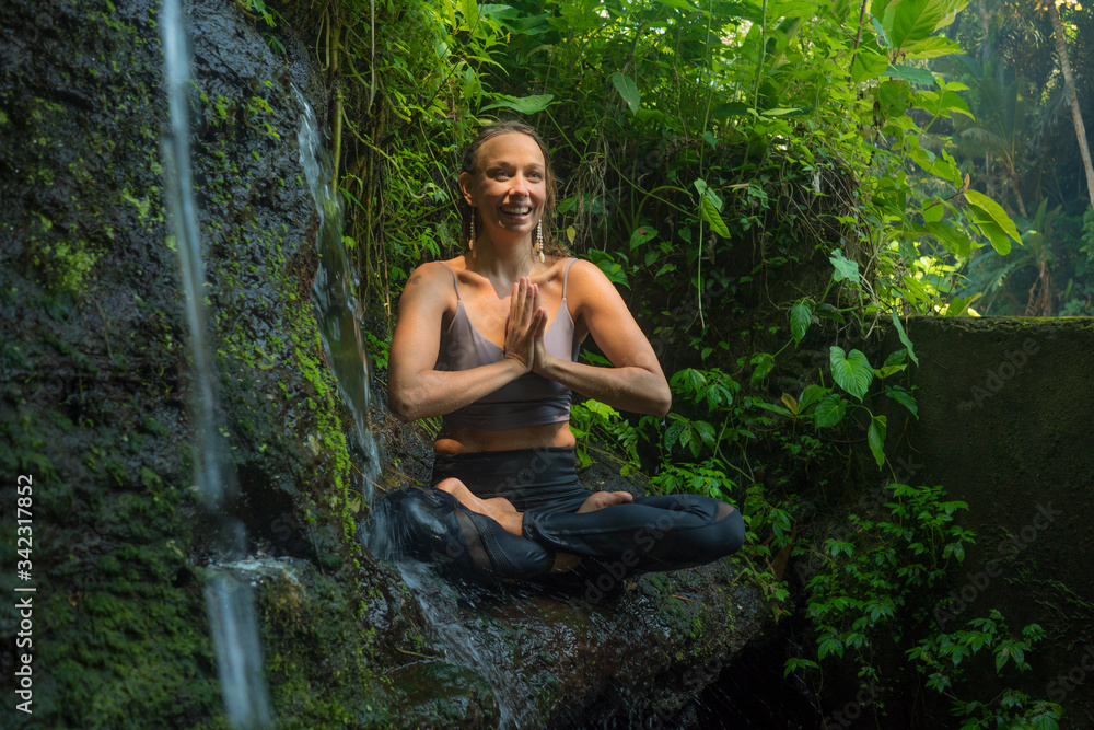Fototapeta premium outdoors adventure portrait of young beautiful and happy woman doing yoga and meditation exercise sitting in lotus posture on rock at tropical rainforest enjoying nature