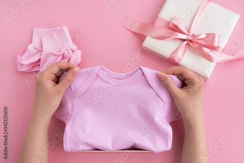 Baby shower concept. Woman hands hold clothes bodysuit. Newborn baby girl accessories on pink background. Top view, flat lay © Elena