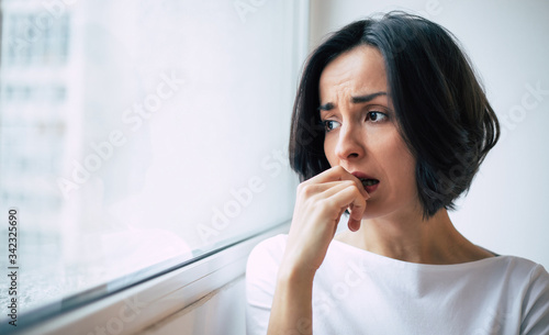 Profound stress. Close-up photo of a sad woman who is looking through the window and biting her nails in distress.