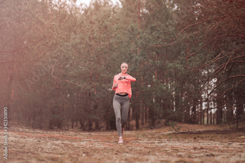 Woman running in the woods and checking smartwatch. Young girl jogging in a forest. Healthy lifestyle fitness sporty woman running early in the morning. Beautiful young woman runner in a pine forest. © ABCreative